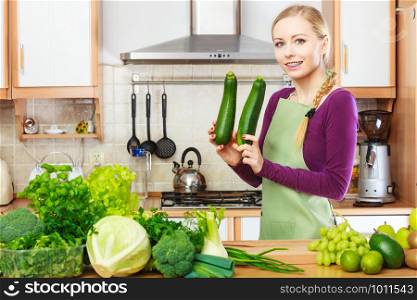 Woman in kitchen with many green leafy vegetables, fresh organically produce on counter. Young happy housewife holding marrow zucchini in hands. Healthy eating, cooking, vegetarian food, dieting and people concept.. Woman housewife in kitchen with green vegetables
