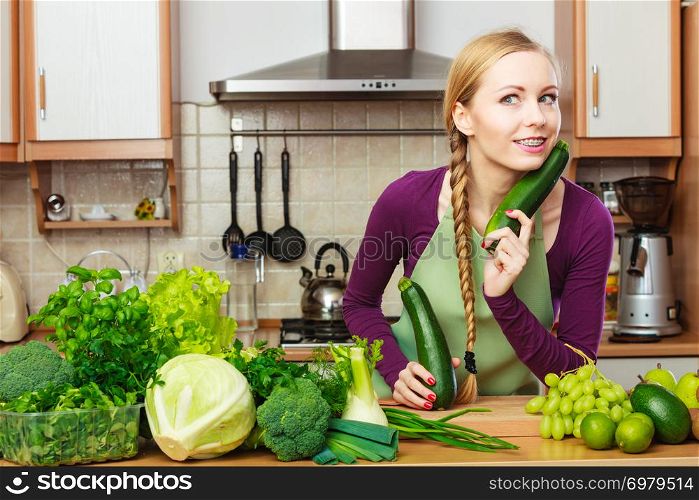 Woman in kitchen with many green leafy vegetables, fresh organically produce on counter. Young happy housewife holding marrow zucchini in hands. Healthy eating, cooking, vegetarian food, dieting and people concept.. Woman housewife in kitchen with green vegetables