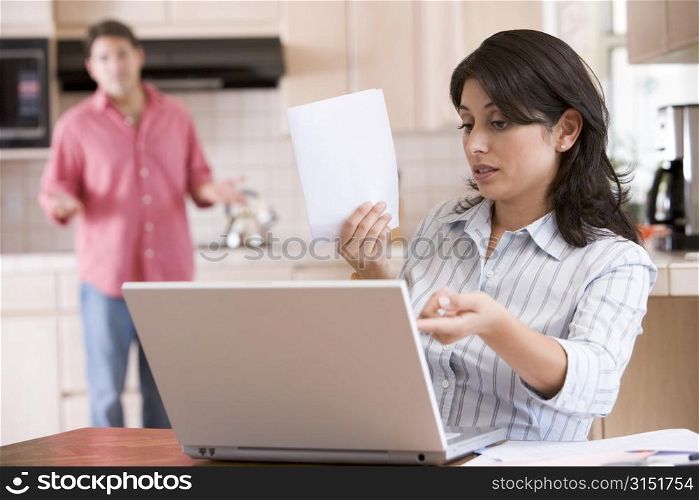 Woman in kitchen with paperwork using laptop with man in background