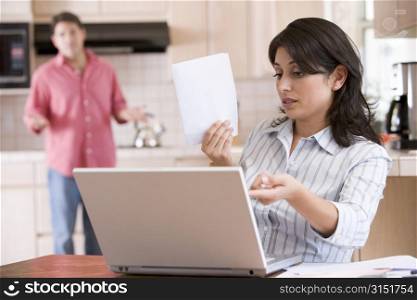 Woman in kitchen with paperwork using laptop with man in background