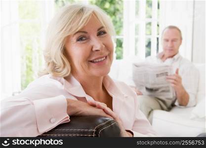 Woman in living room smiling with man in background reading newspaper