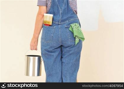 Woman in overalls holding a paint can and brush back pocket - close up