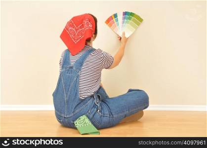 Woman in overalls sitting of wood floor holding paint samples against wall