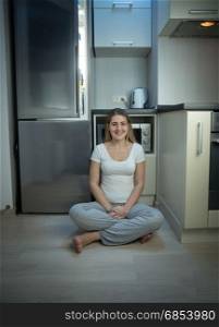 Woman in pajamas sitting on floor at kitchen next to open refrigerator at late evening