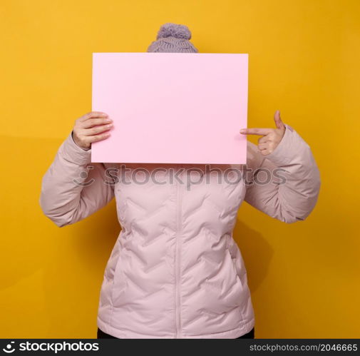 woman in pink winter jacket and hat holds blank pink sheet of paper on yellow background. Seasonal sale. Place for inscription