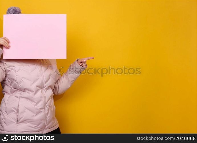 woman in pink winter jacket and hat holds blank pink sheet of paper on yellow background. Seasonal sale. Place for inscription