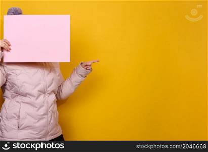 woman in pink winter jacket and hat holds blank pink sheet of paper on yellow background. Seasonal sale. Place for inscription