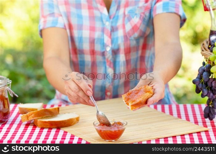 Woman in plaid shirt smears jam on bread with spoon. Red tablecloth, picnic basket, grapes. Sunny day. Picnic concept. Woman in plaid shirt smears jam on bread with spoon