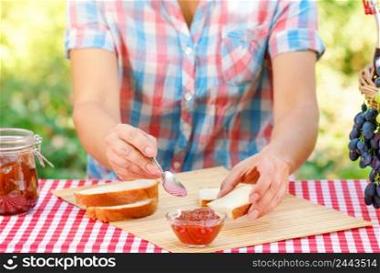 Woman in plaid shirt spreads jam on bread. Red tablecloth, picnic basket, grapes. Sunny day. Picnic concept. Woman in plaid shirt spreads jam on bread
