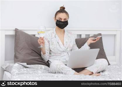 woman in protective mask at home. Young woman using her laptop while relaxing in bed and drinking wine. To stay home. Online study. Remote work. woman in protective mask at home. Young woman using her laptop while relaxing in bed and drinking wine. To stay home. Online study. Remote work.