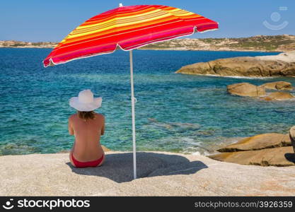 Woman in red bikini and large white floppy hat sitting under a brightly coloured red and yellow striped parasol on a rock and looking out across the blue Mediterranean sea