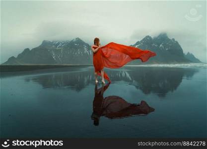 Woman in red dress with waving cape on beach scenic photography. Picture of person with hills on background. High quality wallpaper. Photo concept for ads, travel blog, magazine, article. Woman in red dress with waving cape on beach scenic photography