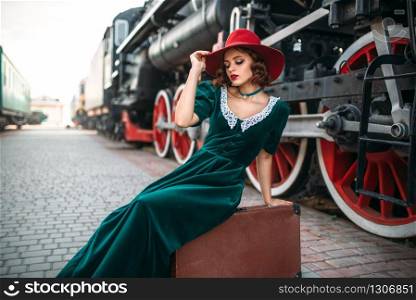 Woman in red hat sitting on suitcase against vintage steam train. Old locomotive. Railway engine, ancient railroad vehicle. Woman in red hat against vintage steam train