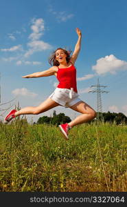 Woman in red t-shirt jumping high on a summer meadow under a blue sky, in the background a power pole is to be seen