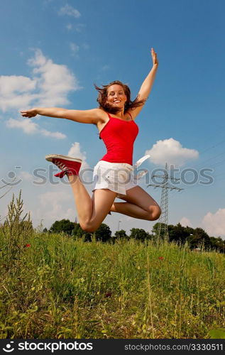 Woman in red t-shirt jumping high on a summer meadow under a blue sky, in the background a power pole is to be seen