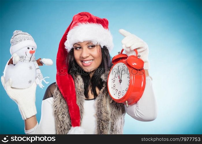 Woman in santa hat with little snowman and clock.. Smiling cute woman in santa claus hat holding little snowman and alarm clock. Pretty mixed race african girl on blue. Christmas season.