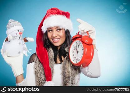 Woman in santa hat with little snowman and clock.. Smiling cute woman in santa claus hat holding little snowman and alarm clock. Pretty mixed race african girl on blue. Christmas season.