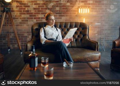 Woman in shirt and trousers reading newspapar with whiskey and cigar, retro fashion, gangster style. Vintage business lady. Woman reading newspapar with whiskey and cigar