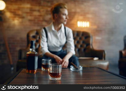 Woman in shirt and trousers sitting at the table with whiskey and cigar, retro fashion, gangster style. Vintage business lady. Woman in shirt and trousers with whiskey and cigar