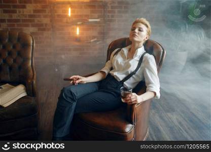 Woman in shirt and trousers sitting in leather chair with whiskey and cigar, retro fashion, gangster style. Woman sitting in chair with whiskey and cigar