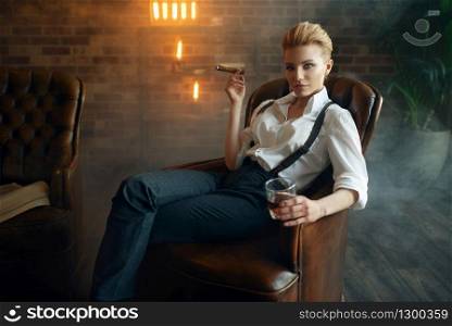 Woman in shirt and trousers sitting in leather chair with whiskey and cigar, retro fashion, gangster style. Vintage business lady in office with brick walls. Woman sitting in chair with whiskey and cigar