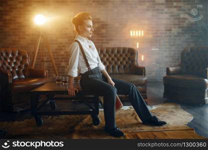 Woman in shirt and trousers with suspenders poses in studio, retro fashion, gangster style. Vintage business lady in office with brick walls. Woman in shirt and trousers, retro fashion