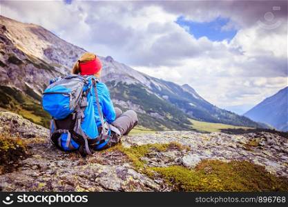Woman in sportswear and with backpack is sitting on the stony ground and enjoys the view, in the mountains