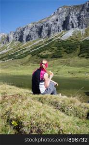 Woman in sportswear is doing a break during a hike: sitting on the ground and enjoying the view over the mountains and a lake