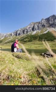 Woman in sportswear is doing a break during a hike: sitting on the ground and enjoying the view over the mountains and a lake