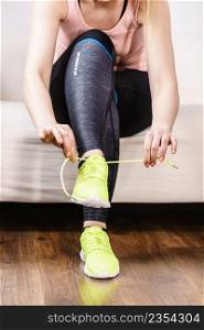 Woman in sportswear sitting on sofa indoor getting ready for exercises. Tying up shoes. Woman putting sport shoes