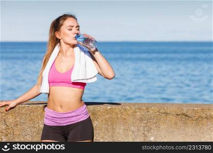 Woman in sportswear takes a break to rehydrate drinking water from plastic bottle, resting after sport workout outdoor by seaside. Woman drinking water after sport gym outdoor