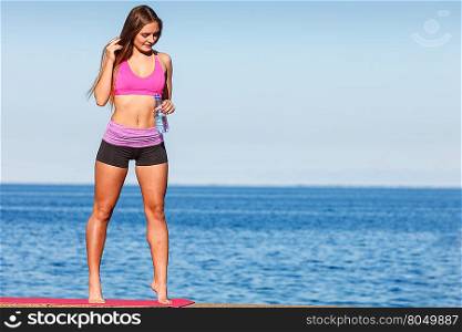 Woman in sportswear takes a break to rehydrate drinking water from plastic bottle, resting after sport workout outdoor by seaside