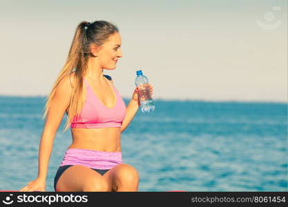 Woman in sportswear takes a break to rehydrate drinking water from plastic bottle, resting after sport workout outdoor by seaside