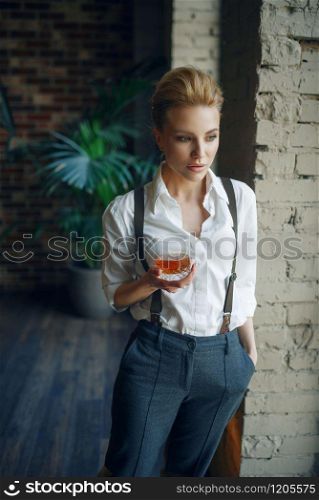 Woman in strict clothes holding glass of whiskey in studio, retro fashion, gangster style. Vintage business lady in office with brick walls. Woman holding glass of whiskey, retro fashion