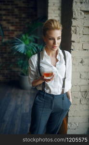 Woman in strict clothes holding glass of whiskey in studio, retro fashion, gangster style. Vintage business lady in office with brick walls. Woman holding glass of whiskey, retro fashion