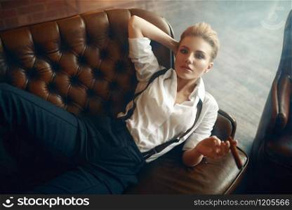 Woman in strict clothes lying on leather couch and smoking cigar, retro fashion, gangster style, female macho. Vintage business lady in office with brick walls. Woman with cigar lying on couch, retro fashion