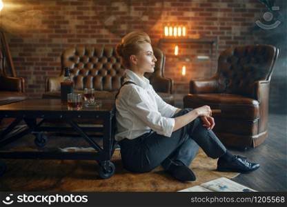 Woman in strict clothes sitting on the floor with whiskey and cigar, retro fashion, gangster style. Vintage business lady in office with brick walls. Woman sitting on the floor with whiskey and cigar