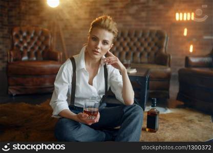 Woman in strict clothes sitting on the floor with whiskey and cigar, retro fashion, gangster style. Vintage business lady in office with brick walls. Woman sitting on the floor with whiskey and cigar