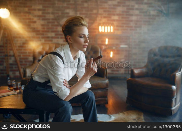 Woman in strict clothes sitting on the table and smoking cigar, retro fashion, gangster style. Vintage business lady in office with brick walls. Woman in strict clothes sitting on the table
