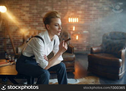 Woman in strict clothes sitting on the table and smoking cigar, retro fashion, gangster style. Vintage business lady in office with brick walls. Woman in strict clothes sitting on the table