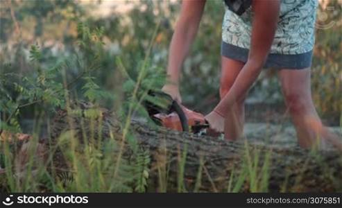 Woman in summer dress and in glasses sawing the trunk of dry wood with chainsaw during summer vacation