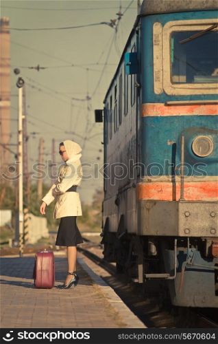 Woman in sunglasses waiting train on railroad station. The autumn cold weather. Beige coat. Retro suitcase.