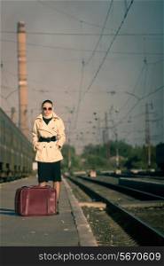 Woman in sunglasses waiting train on railroad station. The autumn cold weather. Beige coat. Retro suitcase.