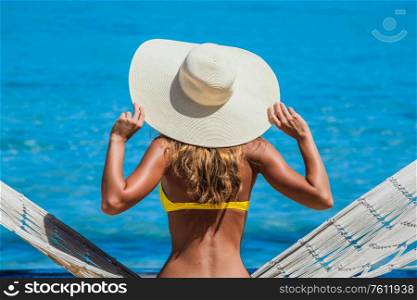 Woman in sunhat swinging in hummock on blue sea background. Woman swinging in hummock