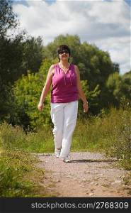 woman in sunny day on wood clearing goes at pathway, in sunglasses