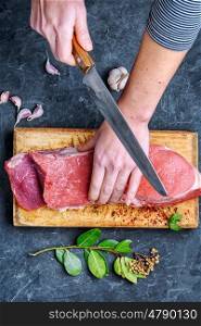 Woman in the kitchen is cutting beef. Man slicing beef stead on wooden cutting board