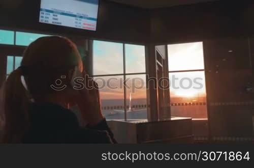 Woman in the terminal of airport talking on the phone and looking out the window with sunset scene