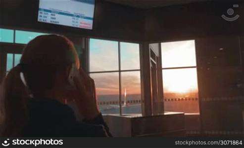 Woman in the terminal of airport talking on the phone and looking out the window with sunset scene