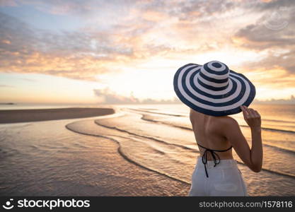 Woman in top bikini and white long pant wearing hat on the beach with a beautiful sunrise or sunset in background.