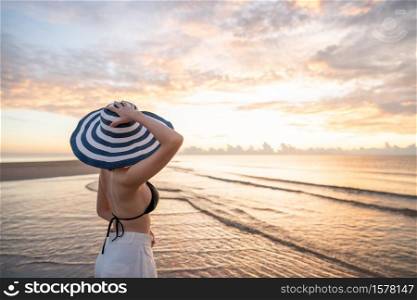 Woman in top bikini and white long pant wearing hat on the beach with a beautiful sunrise or sunset in background.